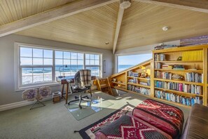 A room with a large window overlooking the ocean, a bookshelf, a desk with a chair, and a patterned rug. - Sanctuary at Seal Rock (Seal Rock)