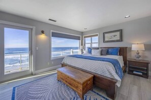 A bedroom with a large bed, a wooden bench, a nightstand, and a view of the ocean. - Sanctuary at Seal Rock (Seal Rock)