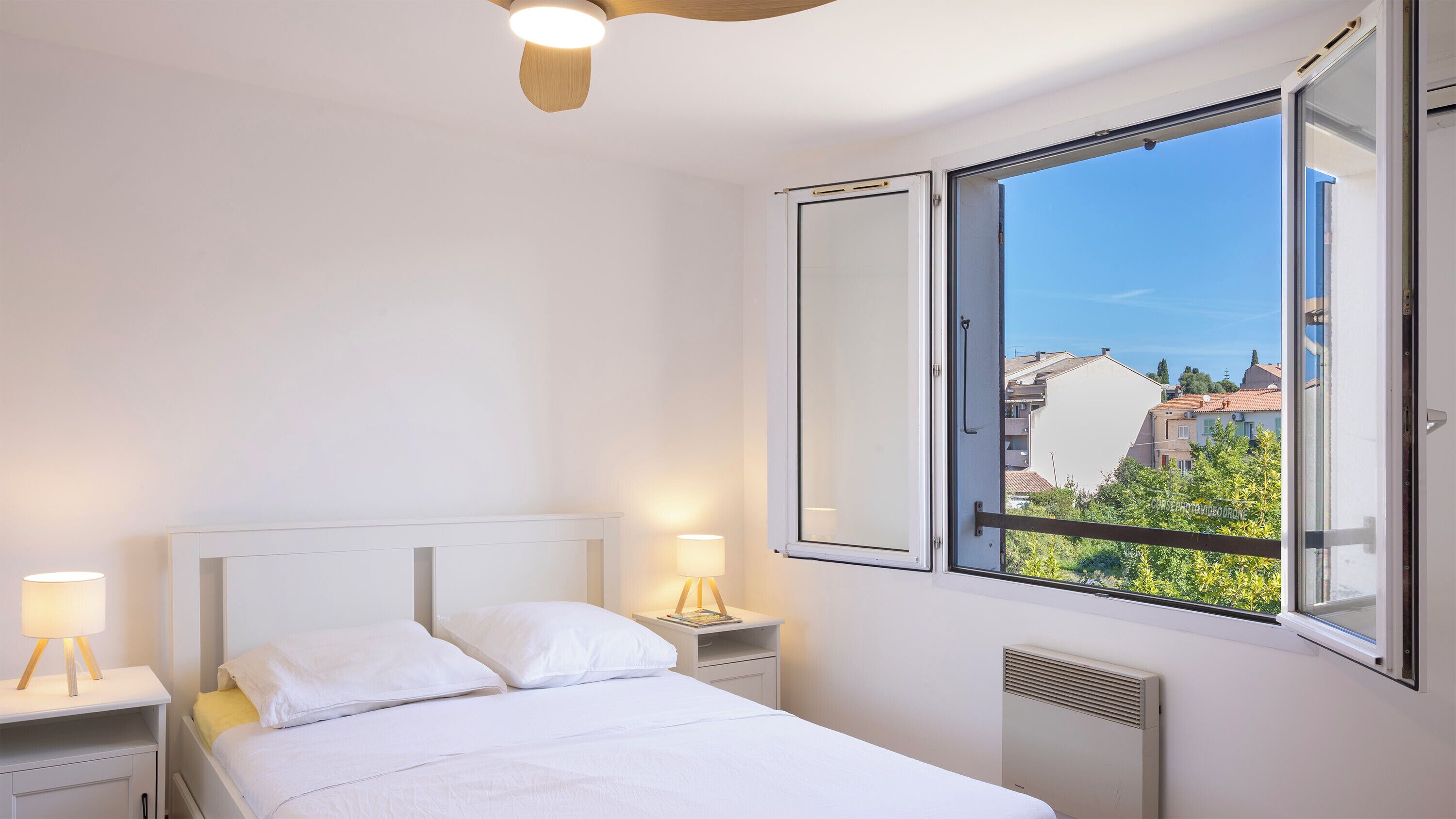 A bedroom with a bed, bedside lamps, a window with an open view, and a ceiling fan.