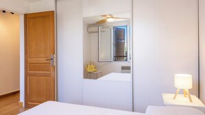 Una camera da letto con una porta in legno, un letto bianco, un comodino con una lampada e uno specchio che riflette una finestra con persiane.