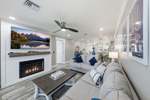 A modern living room with a fireplace, a ceiling fan, and a large landscape painting. - BG North 58 - 2 bed/2Bath - Pool, Tennis, Pickleball, Top Floor - Sleeps 4 (Boca Grande)