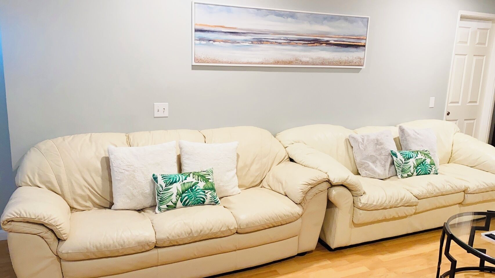 A beige sectional sofa with white and green pillows, a painting above, and a door to the right.