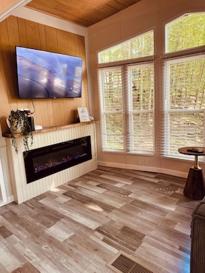 A living room with a flat-screen TV mounted on the wall, a fireplace with a TV screen, and large windows with blinds. - Perfect Nature Getaway Awaits!
Relax, enjoy a peaceful stay surrounded by nature (Milford)