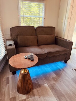 A brown sofa with a round wooden table in front, a window with blinds, and a door with a handle.