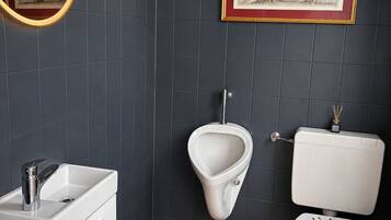 Et moderne badeværelse med et hvidt urinal, et toilet med et dekorativt låg, en vask med armatur og et indrammet kort på væggen.