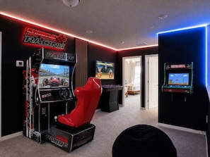 A room with a racing game setup, including a racing simulator chair and two arcade racing machines. - Modern 5BR Windsor Cay Retreat | Private Pool + Game Room (Clermont)