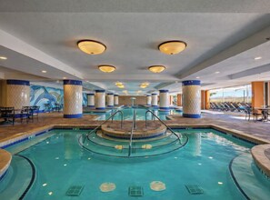 An indoor swimming pool with a central circular platform, surrounded by lounge chairs and a tiled floor. - Oceanfront Condo w/ Pools & Lazy River (North Myrtle Beach)
