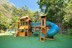 A wooden playhouse with a slide and a swing set in a park.