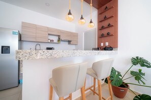 A modern kitchen with a granite countertop, two bar stools, a refrigerator, and a potted plant.