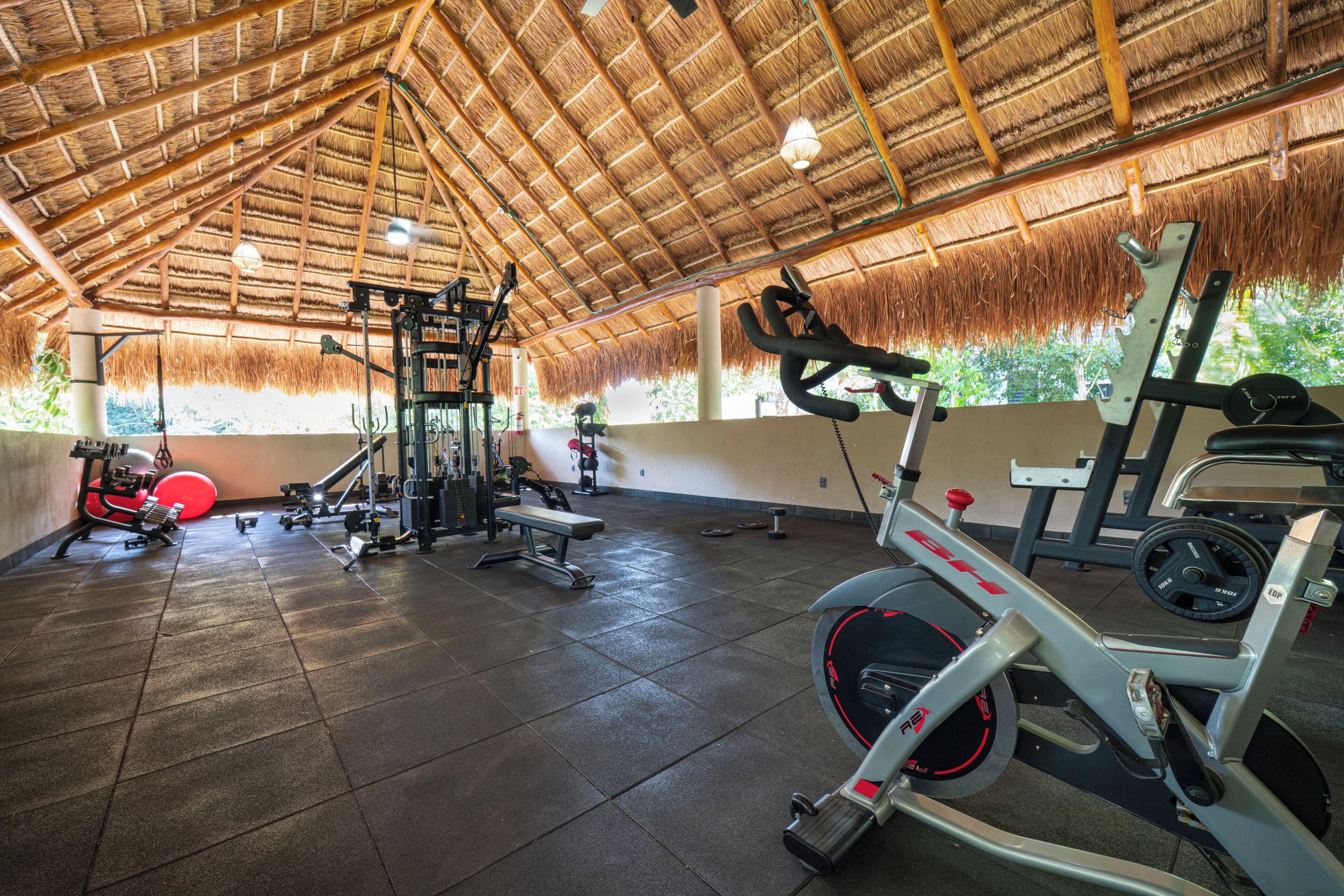 A gym with various exercise equipment, a thatched roof, and a view of greenery outside.