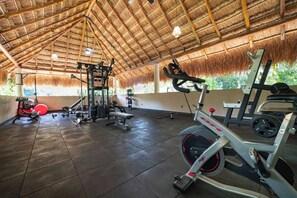 A gym with various exercise equipment, a thatched roof, and a view of greenery outside.