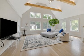 A spacious living room with a large flat-screen TV, a grey sectional sofa, a white armchair, and a large window with a view of greenery. - Waterfront 3 Bedroom Marina Retreat • East Hampton • HeatedPool (East Hampton)