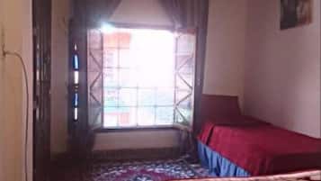 Un dormitorio con cama, una ventana con cortinas, un tapete estampado y una lámpara fijada a la pared.