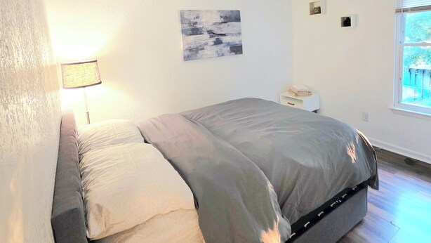 Un dormitorio con cama, mesita de noche, lámpara, ventana y una obra de arte colgada en la pared.