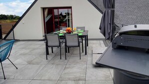Uma área de refeições moderna ao ar livre, com mesa posta para dois, churrasqueira e área de estar coberta.