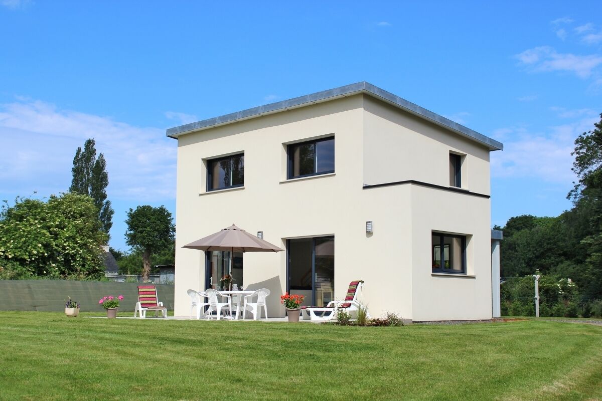 A modern two-story building with a flat roof, large windows, and a patio area with outdoor furniture.