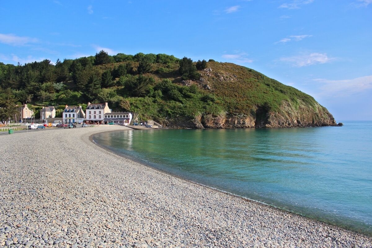 A pebble beach with a curved shoreline, buildings on the left, and a hill with greenery in the background.
