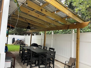 Eine Terrasse im Freien mit einer Holz-Pergola, einem Esstisch mit Stühlen und einem weißen Zaun im Hintergrund.