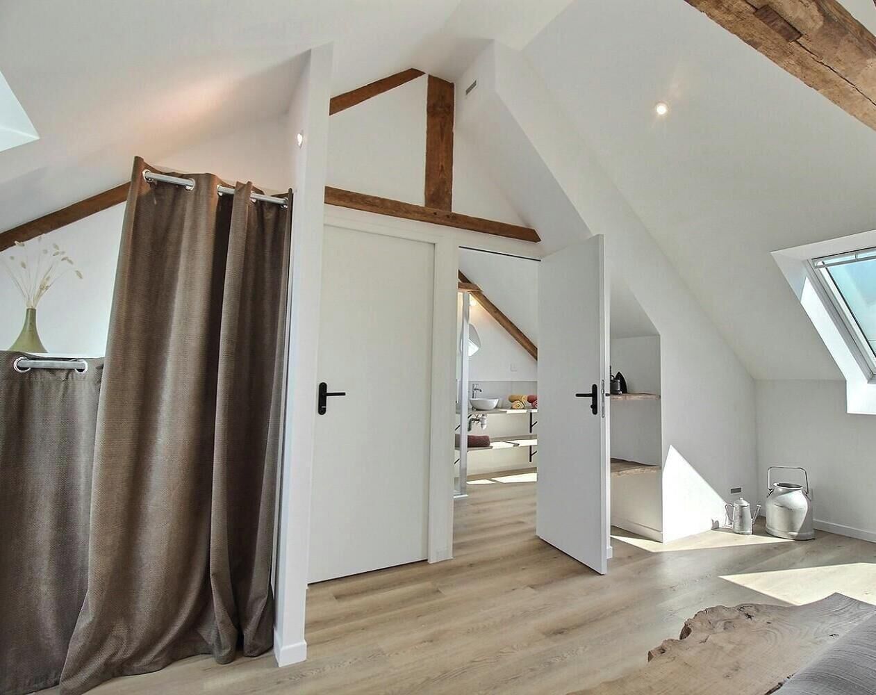 Une chambre située dans un espace mansardé, avec un plafond en pente, des poutres apparentes, une lucarne et une vue sur la cuisine.