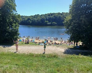Ein Strand am See mit Badegästen und Sonnenanbetern, umgeben von Bäumen.