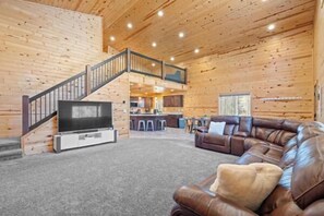 A spacious living room with a leather couch, a flat-screen TV, and a staircase leading to an upper level. - Steelhead-Modern Yellowstone Cabin, Fiber Wi-Fi & Pine Views & Air Conditioning! (Island Park)