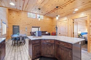 A wooden interior with a kitchen area featuring a large island, dark wooden cabinets, and a dining table with chairs. - Steelhead-Modern Yellowstone Cabin, Fiber Wi-Fi & Pine Views & Air Conditioning! (Island Park)