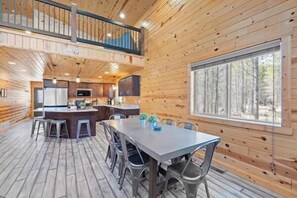 A cabin interior with a dining area, kitchen, and a large window. - Steelhead-Modern Yellowstone Cabin, Fiber Wi-Fi & Pine Views & Air Conditioning! (Island Park)
