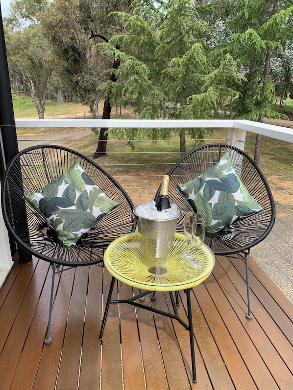 Una terraza de madera con dos sillas de alambre negras, cada una con un cojín estampado con motivos de hojas, un balde de hielo metálico y un vaso sobre una mesa redonda amarilla.