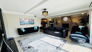 A living room with a black sofa, a patterned rug, a fireplace, and a clock on the wall. - Coastal Garden: 1 Block to Stewart Beach, 4BR Sleeps 12, Luxury Bidets (Galveston)