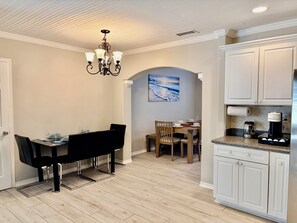 A dining area with a table set for four, a chandelier, and a painting of a beach scene on the wall. - Coastal Garden: 1 Block to Stewart Beach, 4BR Sleeps 12, Luxury Bidets (Galveston)