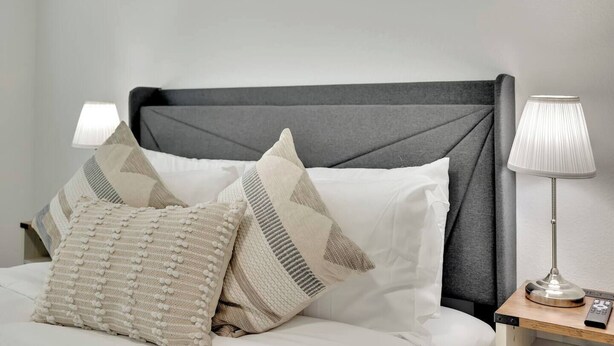 A neatly made bed with a grey headboard, multiple pillows, and a bedside lamp on a wooden nightstand. - Modern 5B3B｜Near Strip｜Fiber WiFi｜TV in all Room (Las Vegas)