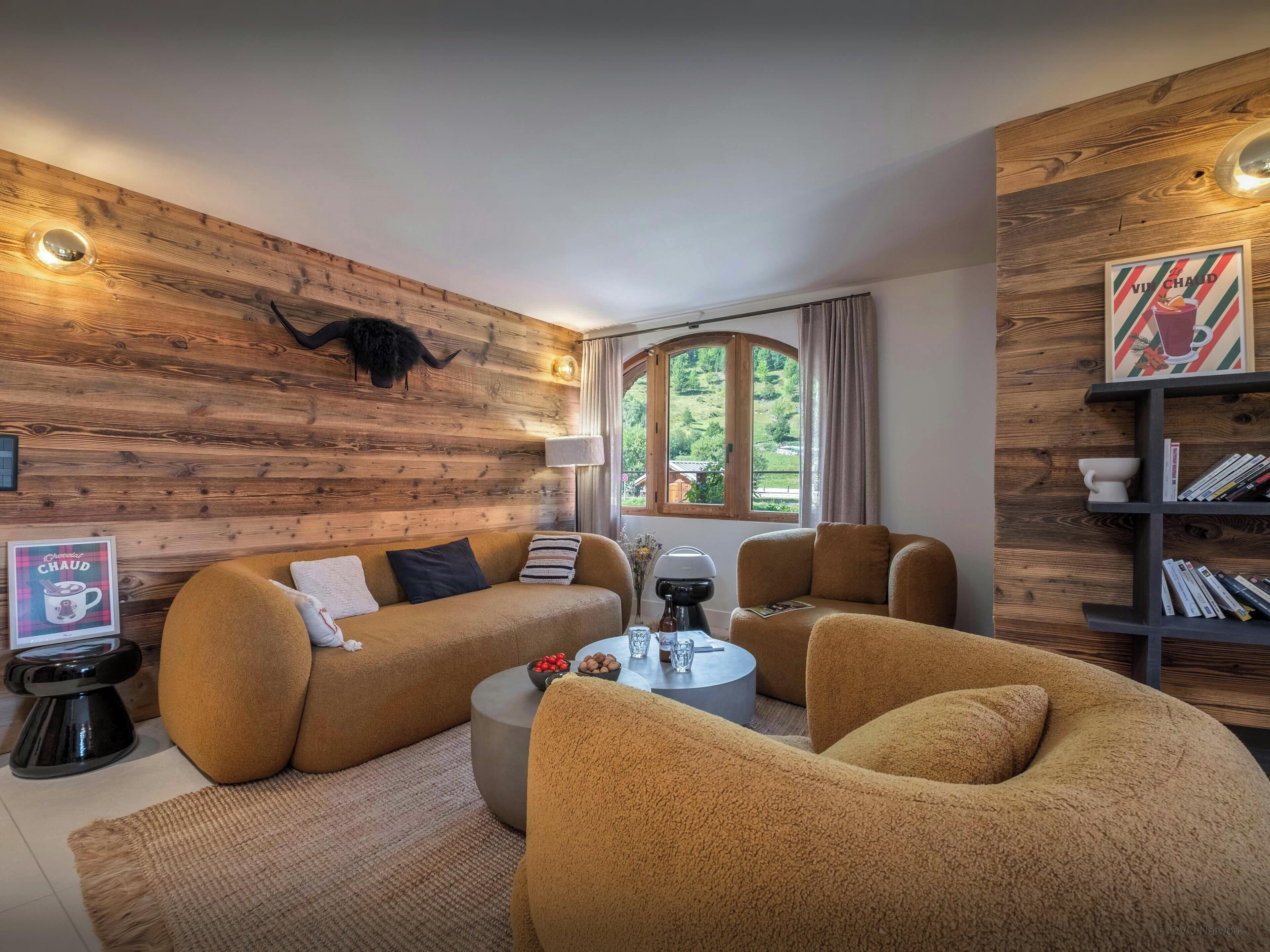 A cozy living room with a wooden wall, a sofa, armchairs, a coffee table, and a bookshelf.