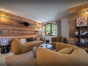 A cozy living room with a wooden wall, a sofa, armchairs, a coffee table, and a bookshelf.