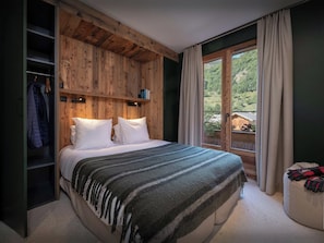 A bedroom with a wooden headboard, a bed with white bedding, a window with curtains, and a view of greenery outside.