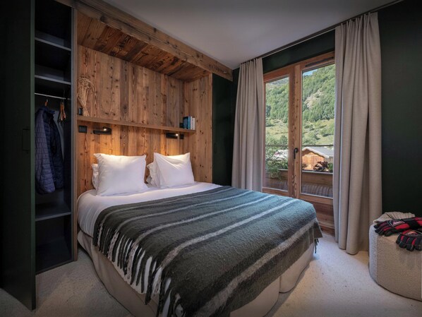 A bedroom with a wooden headboard, a bed with white bedding, a window with curtains, and a view of greenery outside.