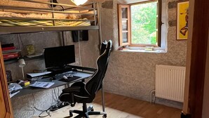 Quarto pequeno com uma cama de solteiro, uma escrivaninha com computador, uma cadeira e uma janela com vista para o verde.