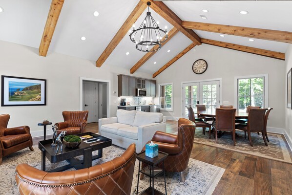 A spacious living room with leather furniture, a chandelier, and a view of the kitchen.