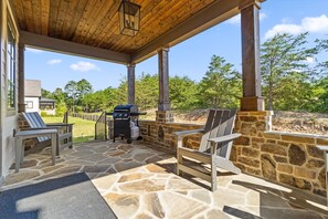 A covered outdoor patio with stone flooring, two Adirondack chairs, a grill, and a view of a house and trees.
