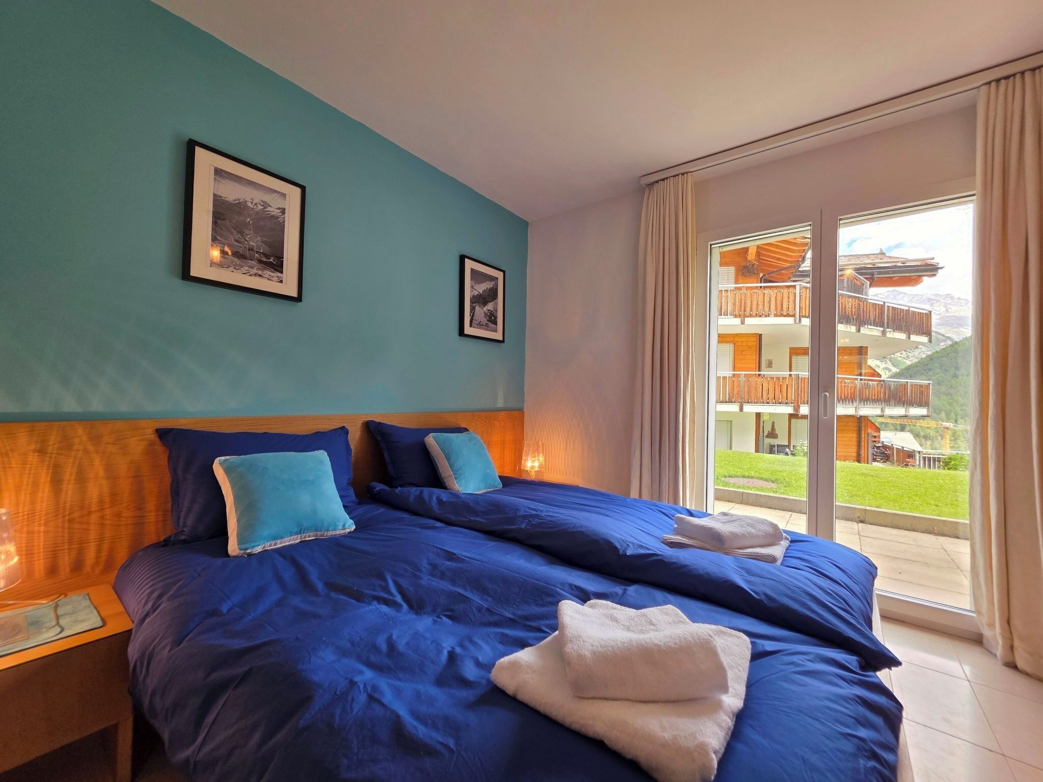 Ein Hotelzimmer mit einem großen Bett, zwei gerahmten Bildern an der Wand und Blick auf ein Gebäude mit Balkonen durch eine Glastür.