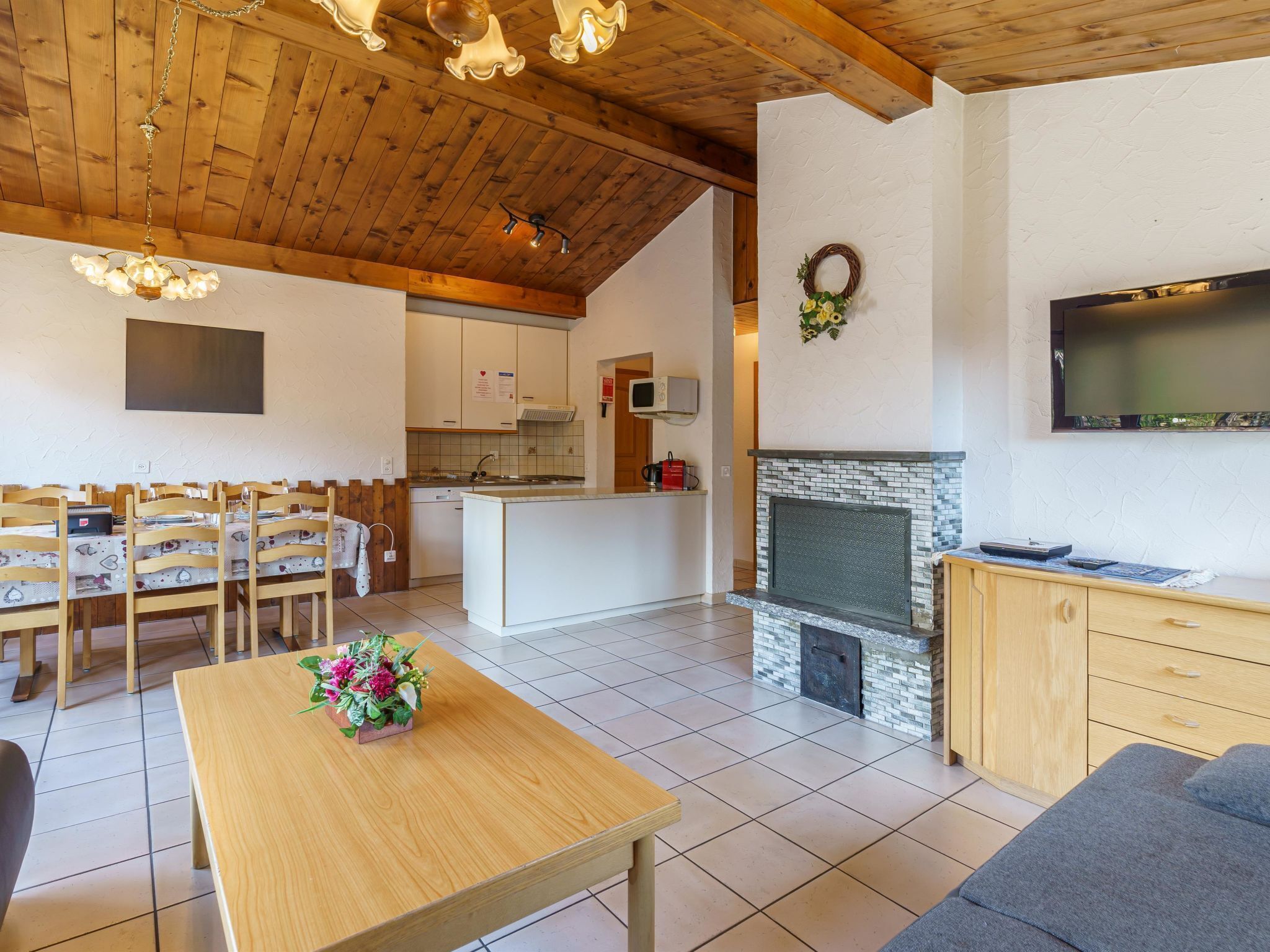 A spacious living area with a wooden ceiling, a fireplace, a kitchen in the background, and a dining area with wooden furniture.