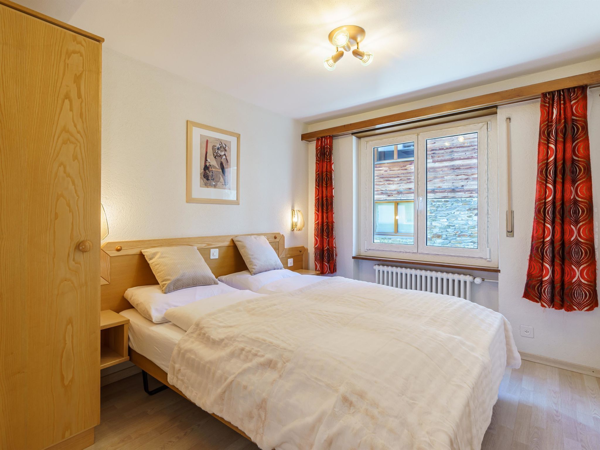 Ein Schlafzimmer mit einem Bett, einem Nachttisch, einer Wandlampe, einem Fenster mit Vorhängen und einer Heizung.