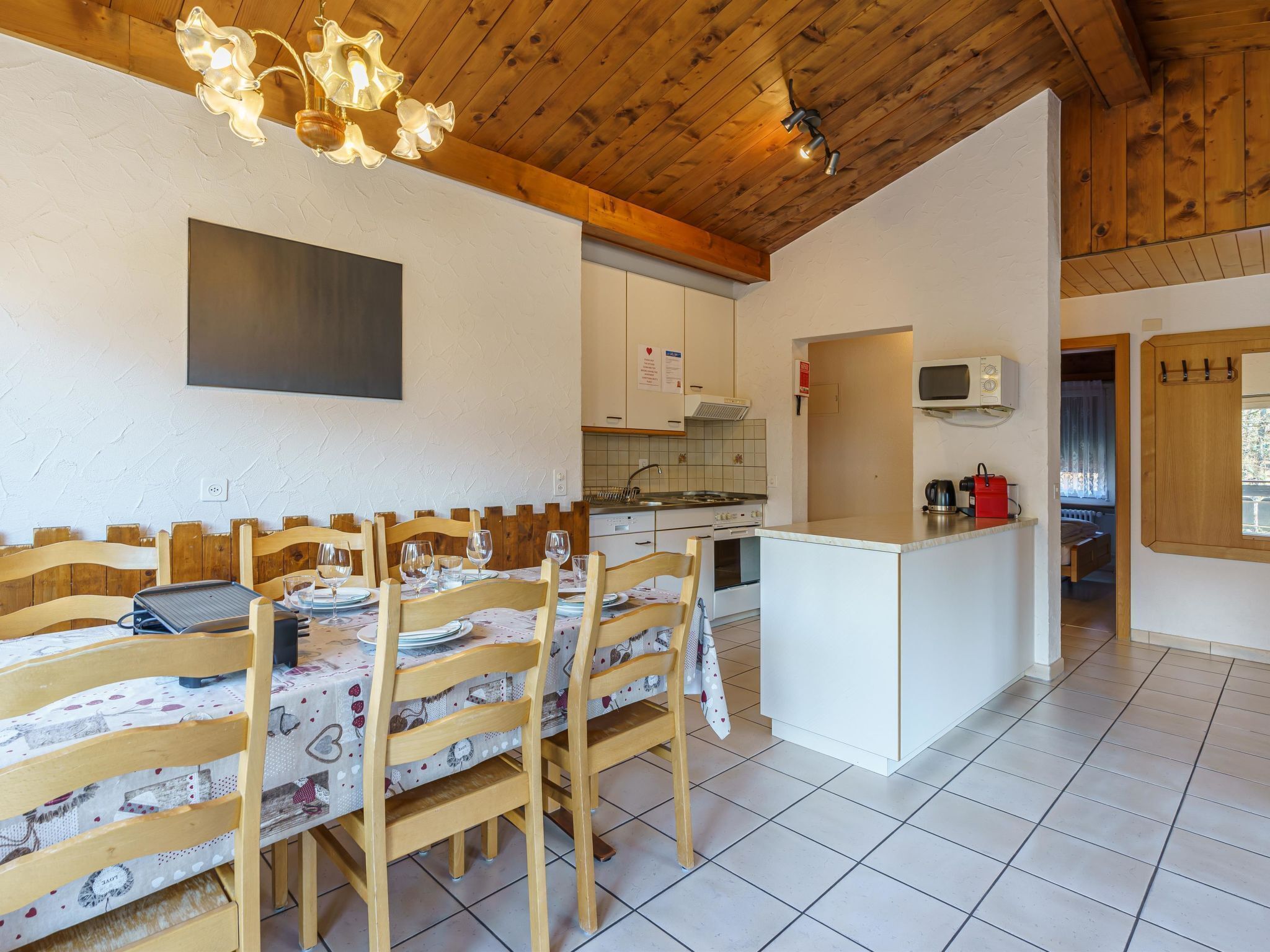 A dining area with wooden chairs and a table set for four, a kitchen with white cabinets and a microwave, and a wooden ceiling with exposed beams.