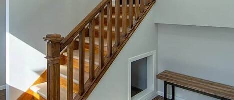 Una escalera de madera con barandilla, un pequeño banco y un tapete estampado sobre piso de baldosas.