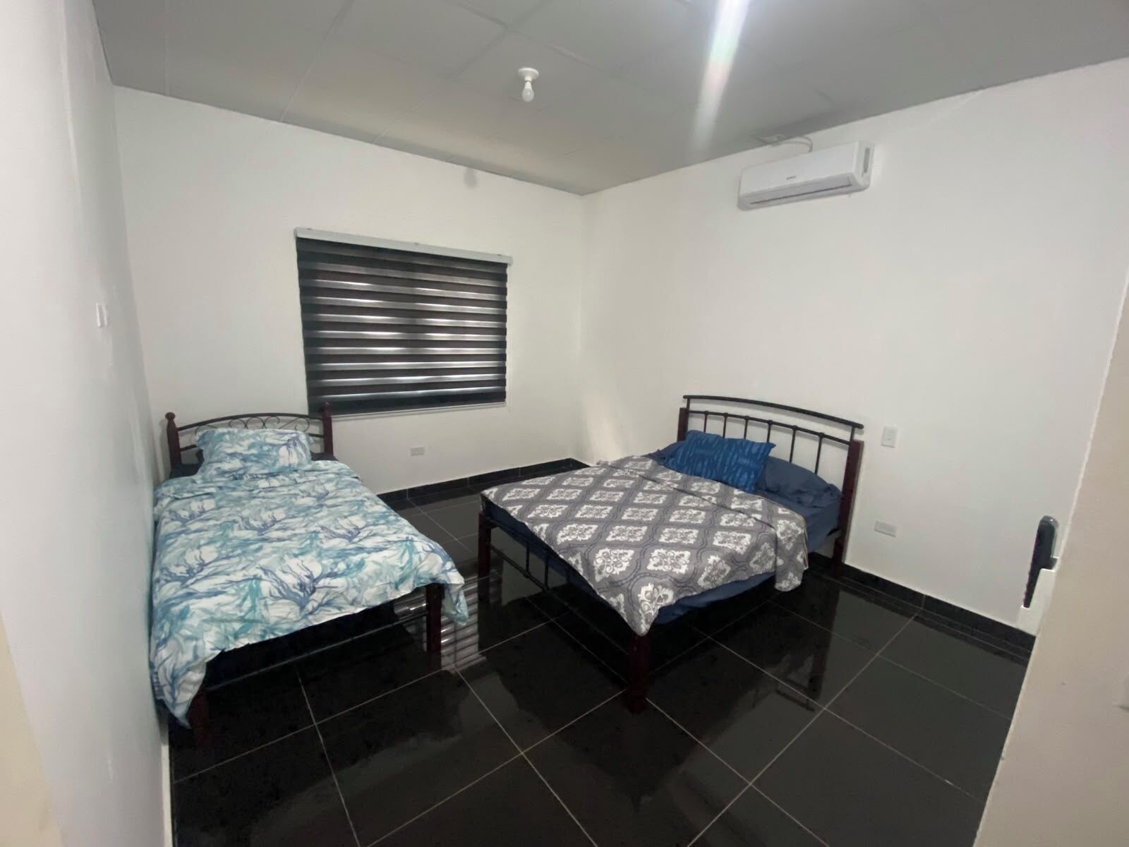 A small, clean room with two single beds, a window with blinds, and an air conditioning unit on the wall.