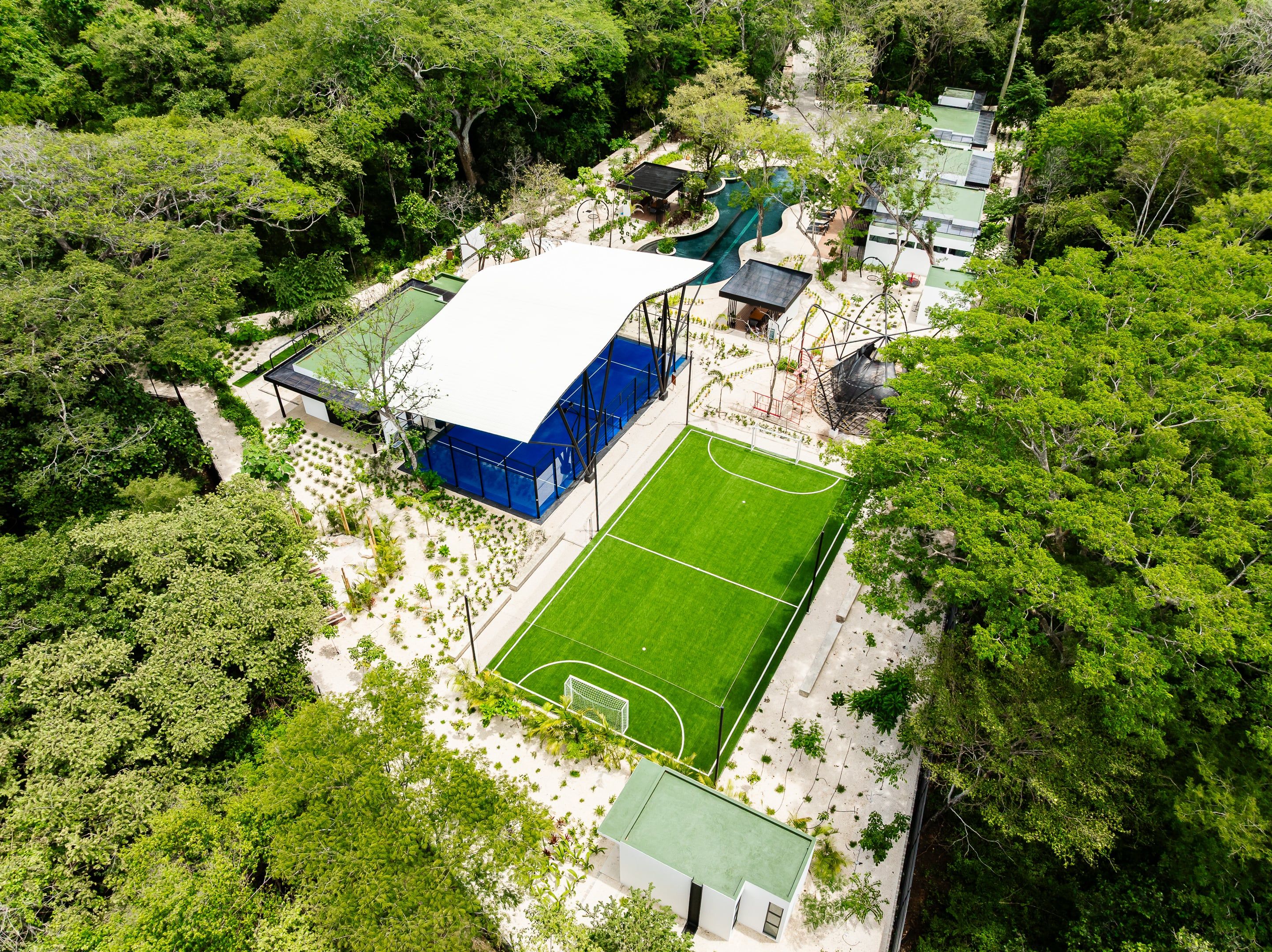 An aerial view of a property with a large building, a tennis court, and a pool area surrounded by dense greenery.