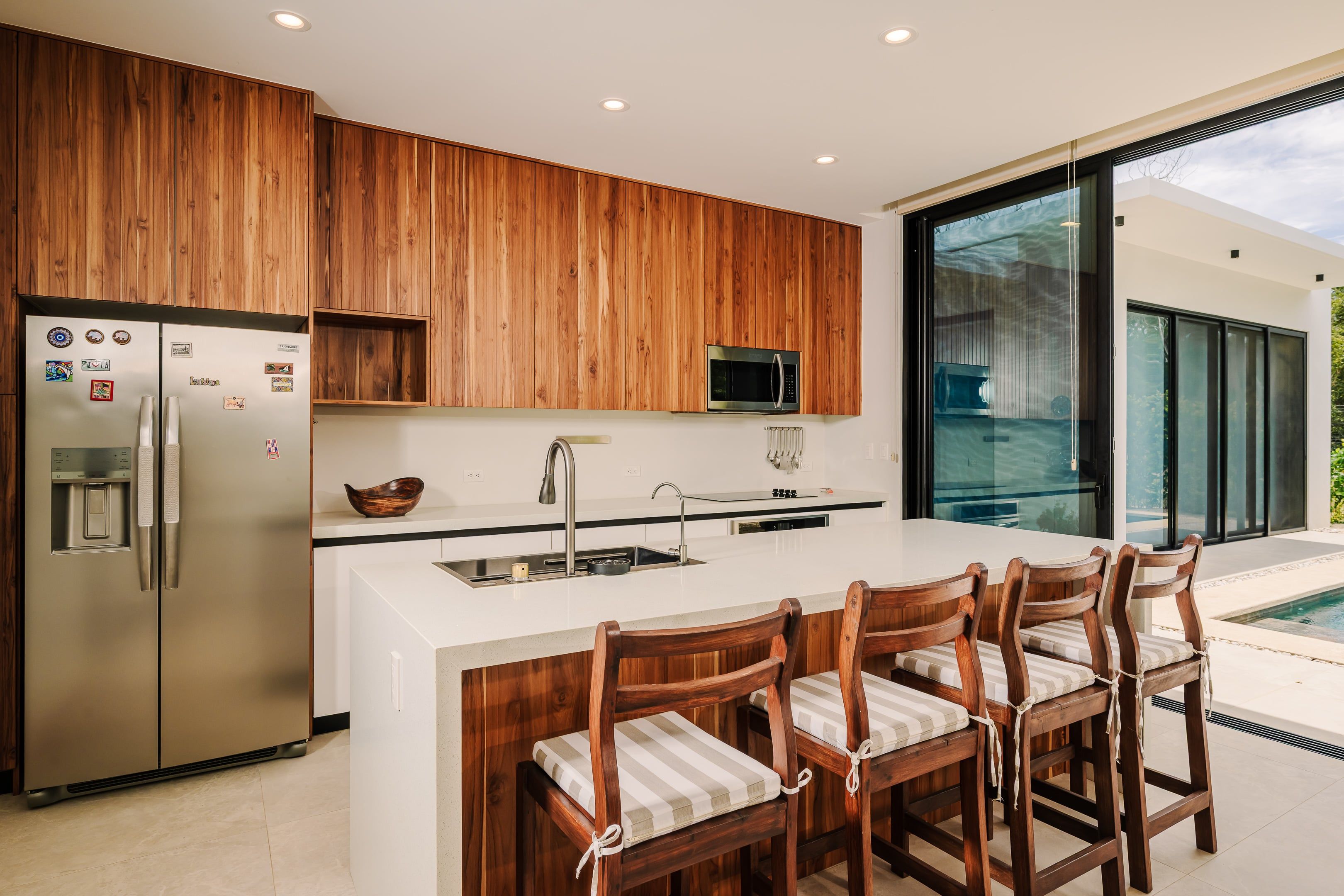 A modern kitchen with wooden cabinets, stainless steel appliances, a central island with bar stools, and a large window offering a view of the outdoors.