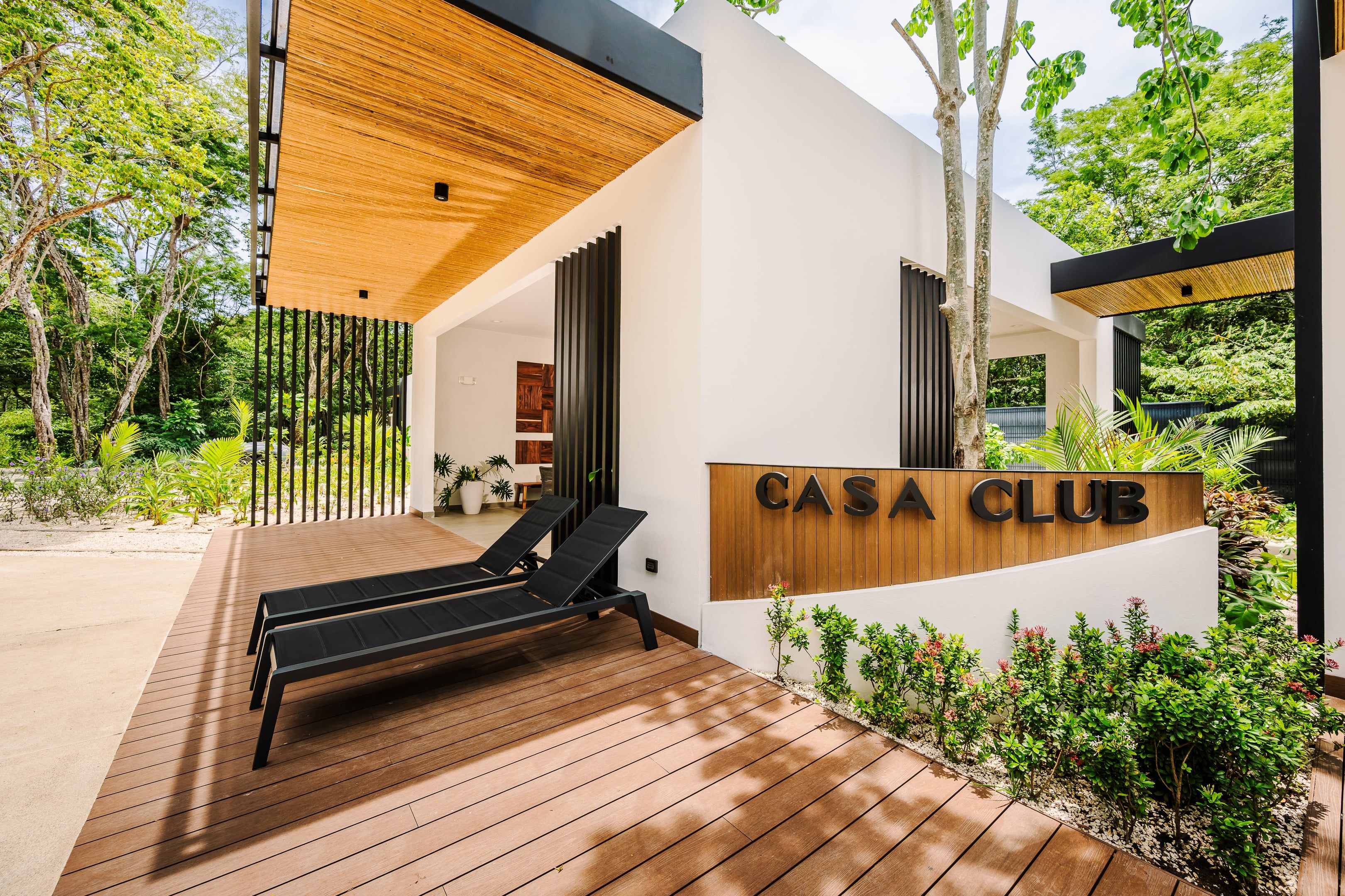 A modern outdoor lounge area with wooden flooring and black lounge chairs, a wooden pergola, and a sign reading 'CASA CLUB'.