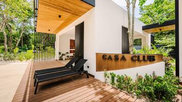 A modern outdoor lounge area with wooden flooring and black lounge chairs, a wooden pergola, and a sign reading 'CASA CLUB'.