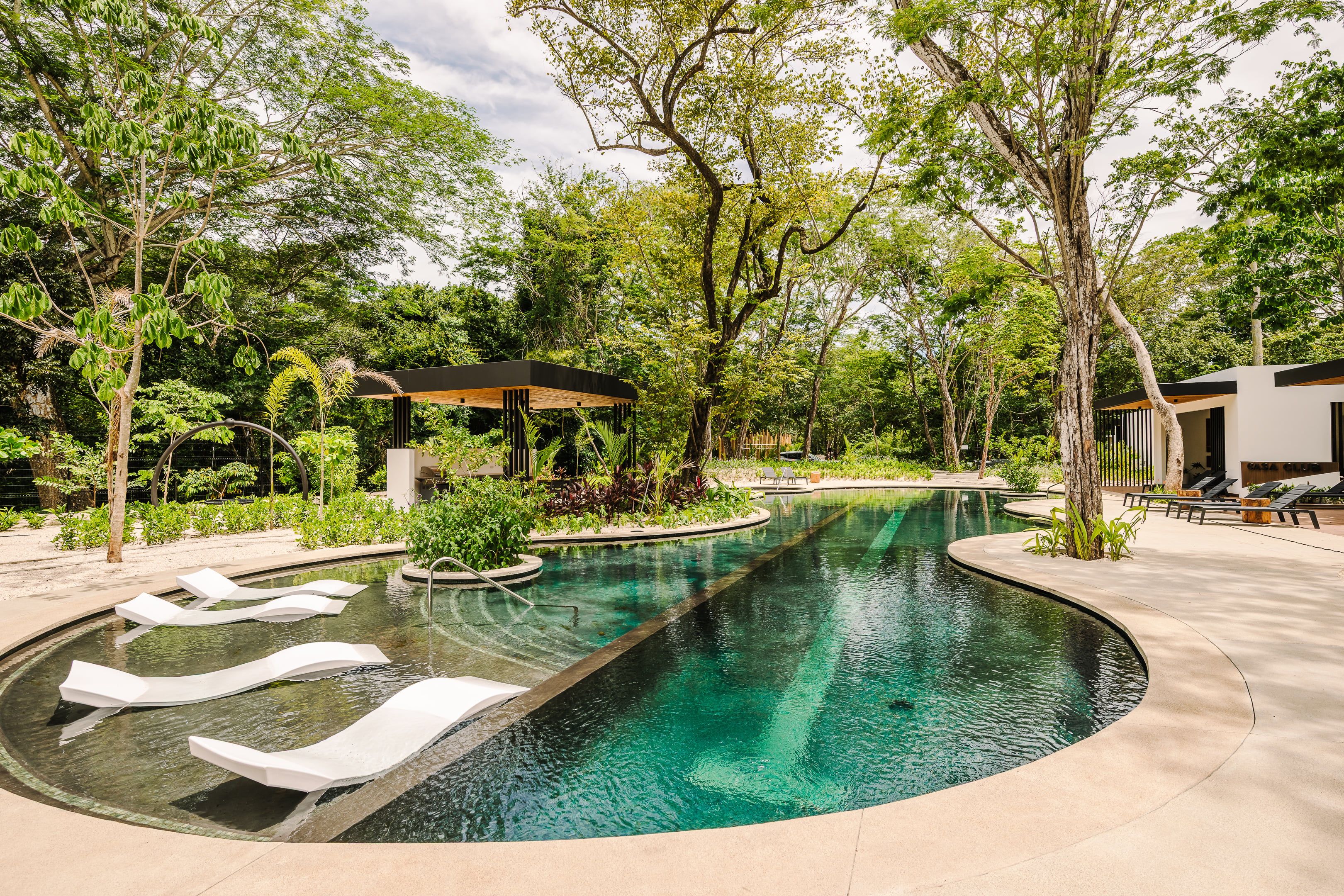 A pool with a curved design, surrounded by lush greenery and a gazebo.