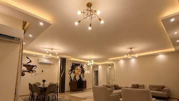 A modern living room with a dining area, featuring a central chandelier, sofas, and a coffee-themed wall art.